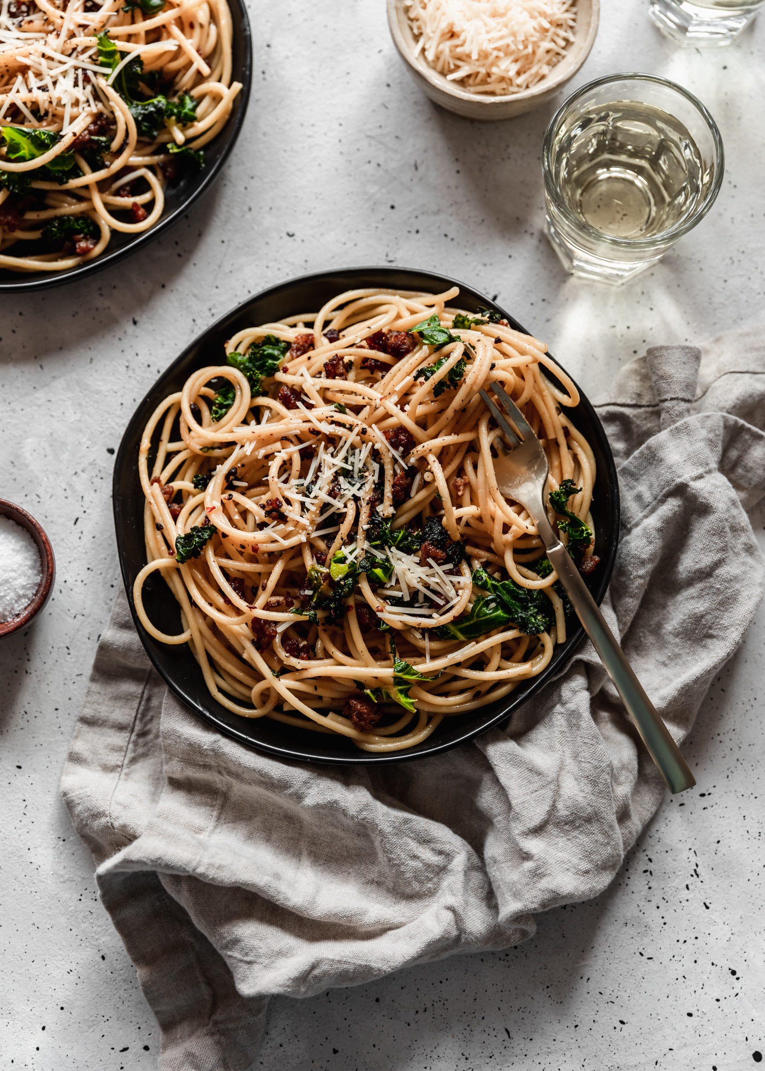 An overhead image of a black plate with bucatini carbonara, sausage, and kale placed on a grey linen on a grey speckled table surrounded by another plate of pasta, wine, and a small wood bowl of salt.