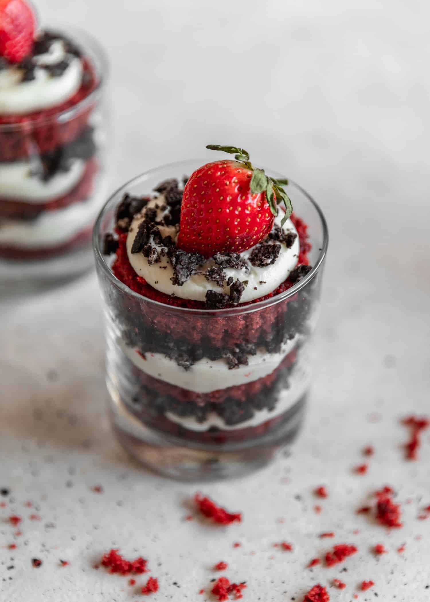 A closeup 45 degree photo of two red velvet trifles on a grey table surrounded by cake crumbs.