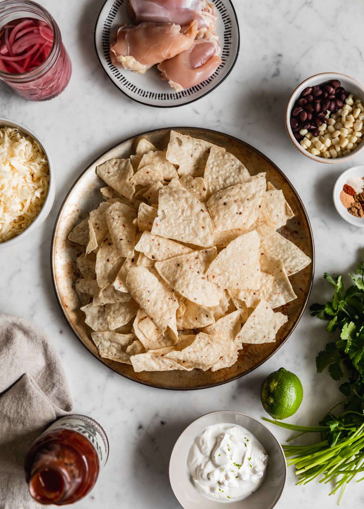 An overhead picture of a plate of tortilla chips surrounded by meat, veggies, cheese, sour cream, and spices on a marble counter.