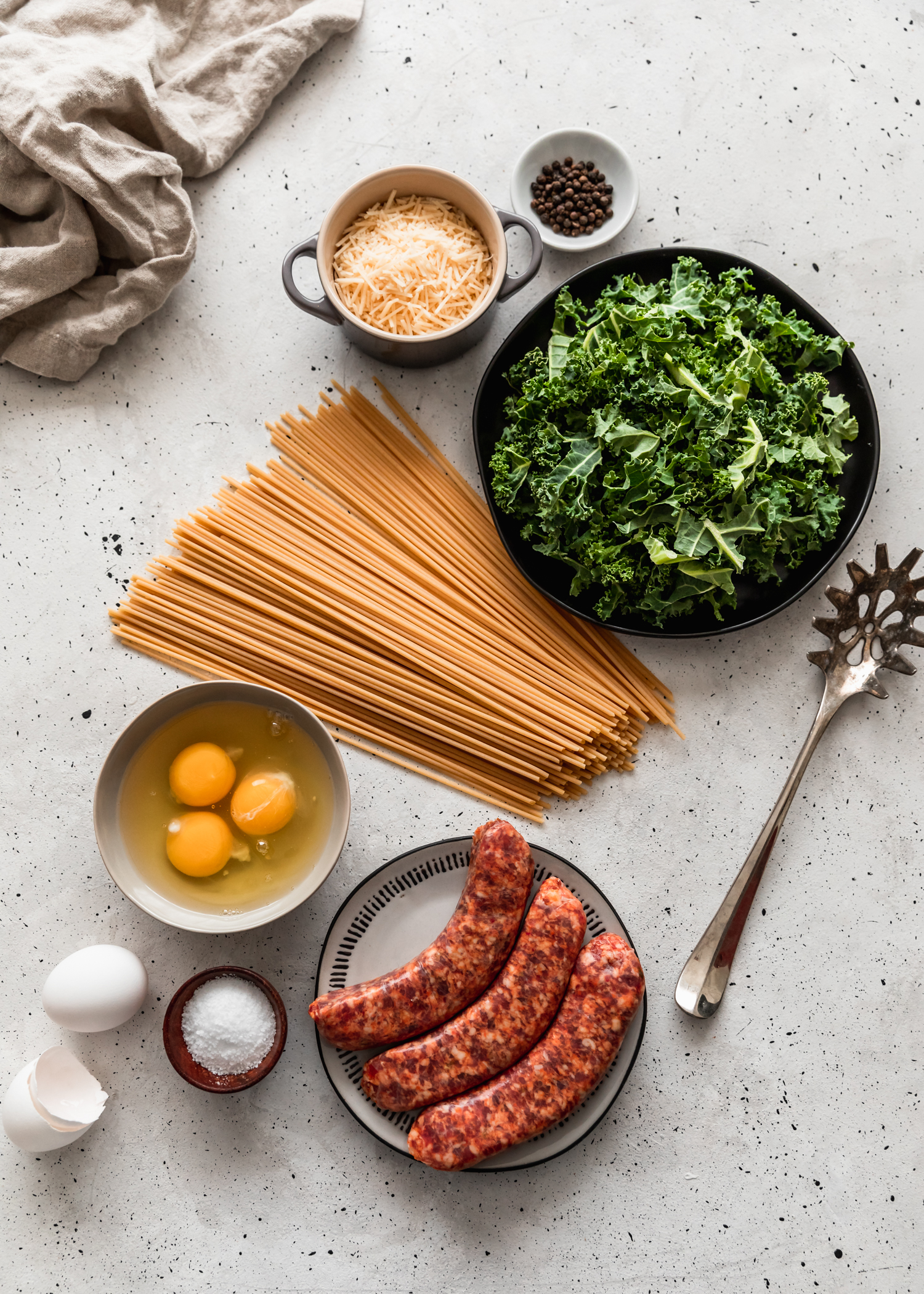 An overhead picture of bucatini on a grey table surrounded by a plate plate of kale, a grey bowl of parmesan, a white pinch pot with peppercorns, a grey bowl with eggs, and a black and white plate topped with sausage.