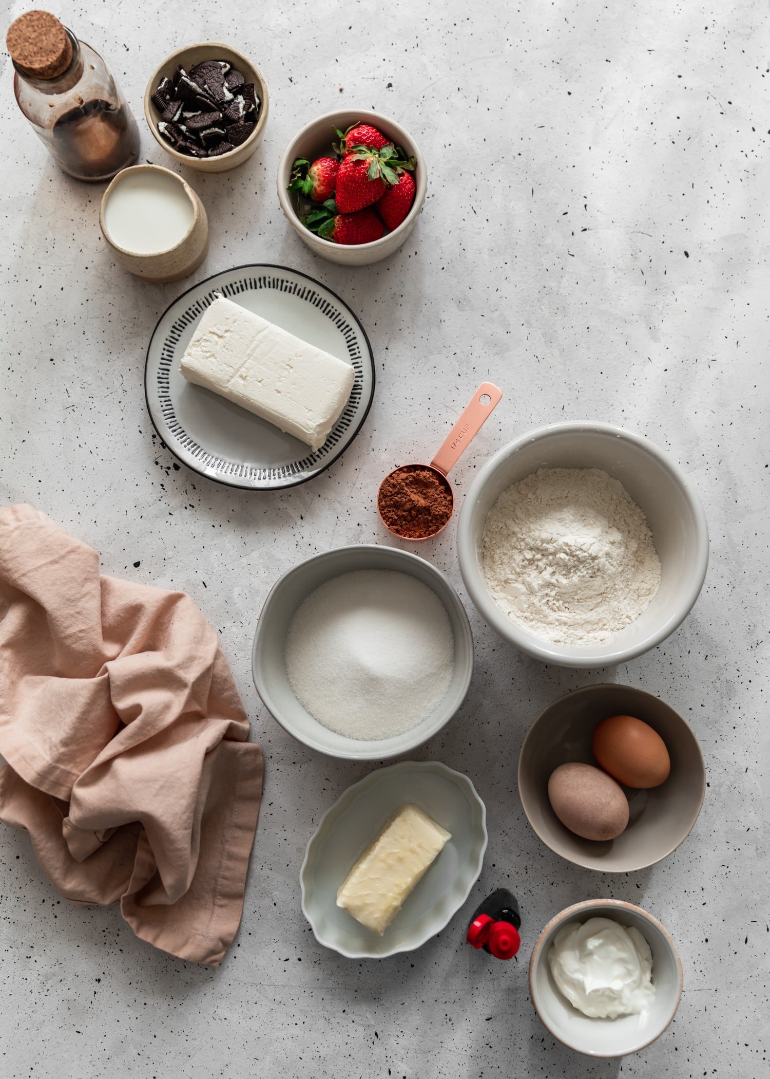 An overhead image of various grey and white bowls filled with cake and frosting ingredients on a grey speckled table next to a pink linen.