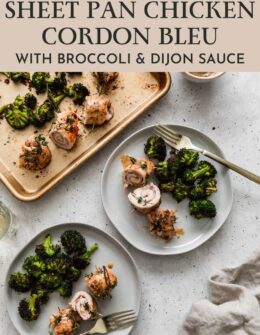 An overhead image of two white plates with chicken cordon bleu rolls and broccoli on a white speckled table next to a gold sheet pan with more food, a beige linen, and a white bowl of Dijon sauce.