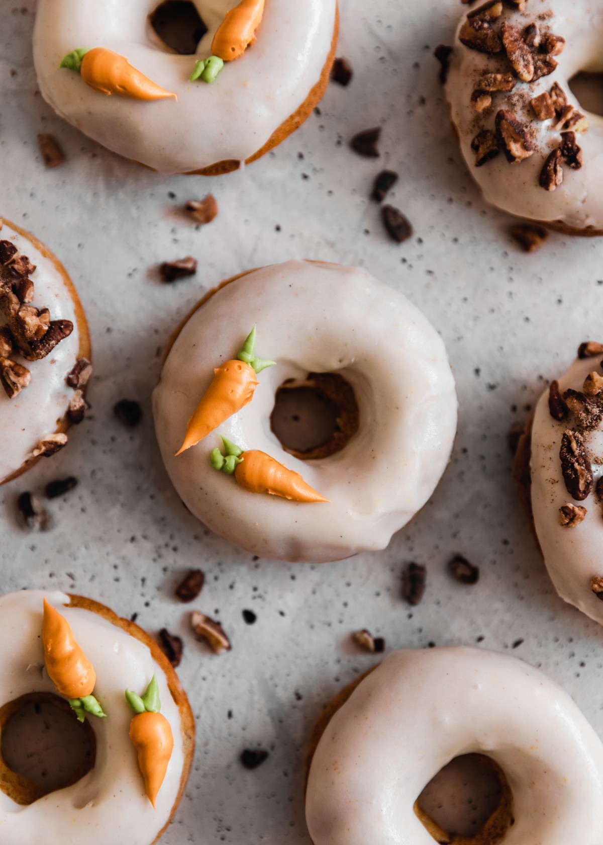 On overhead image of carrot cake donuts with white glaze, decorated with frosting carrots and pecans on a grey speckled table.