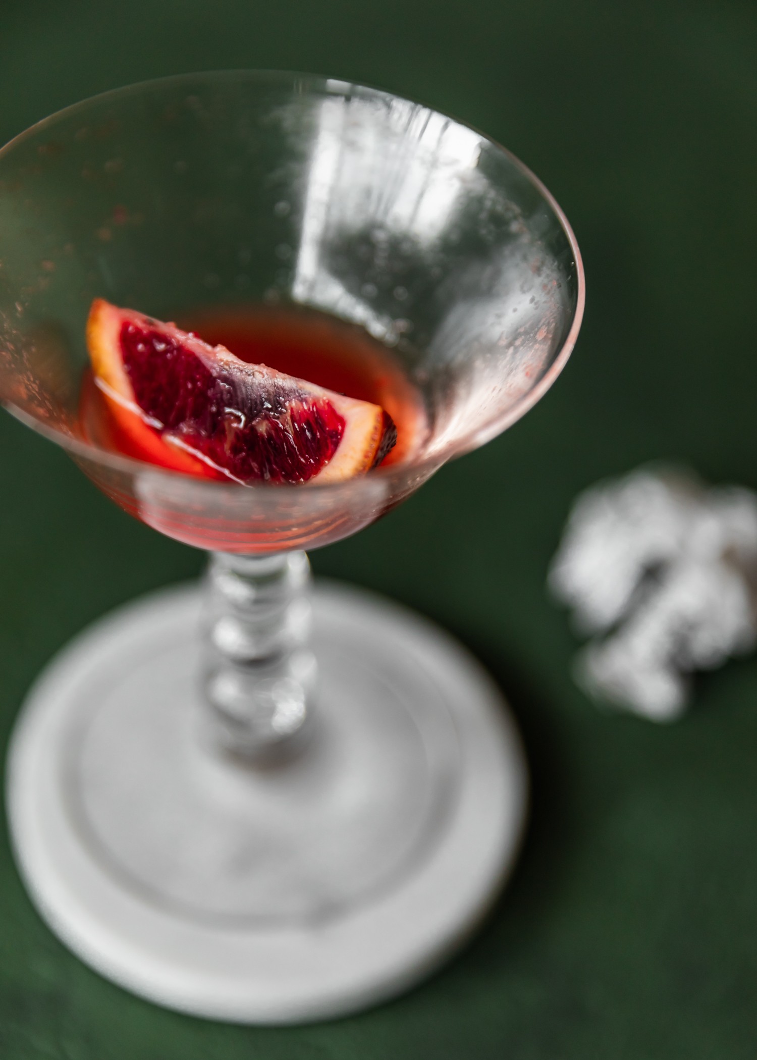 An extremely closeup picture of an almost-empty coupe glass with a red cocktail, focused on a slice of blood orange. The glass is on a marble coaster on a dark green table next to a crumpled piece of paper.