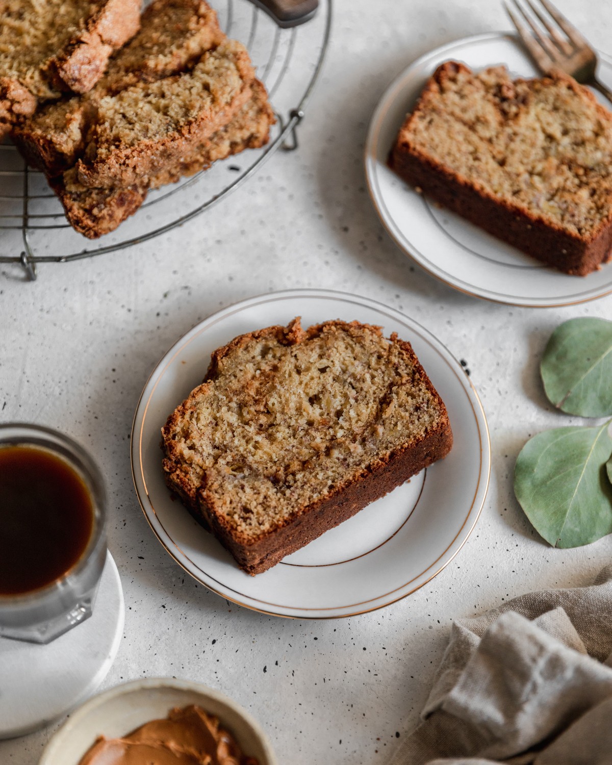A side image of a slice of banana bread on a white plate placed on a white counter surrounded by cups of coffee, plates, a beige linen, and greenery.