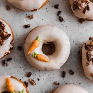 A bird's eye photo of a donut with frosting carrots on a grey table surrounded by more donuts and pecans.