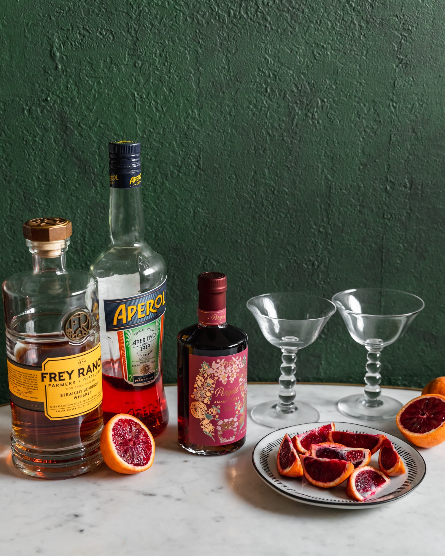 A side image of a bottle of bourbon, Aperol, and a bottle of Amaro on a marble counter surrounded by sliced blood oranges and two coupe glasses with a dark green background.