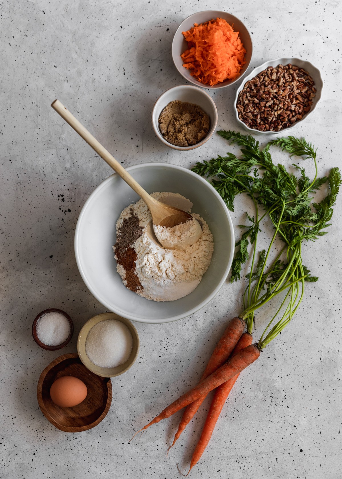 Ingredients for carrot cake donuts, including sugar, pecans, carrots, and dry goods in white and beige bowls on a grey counter.