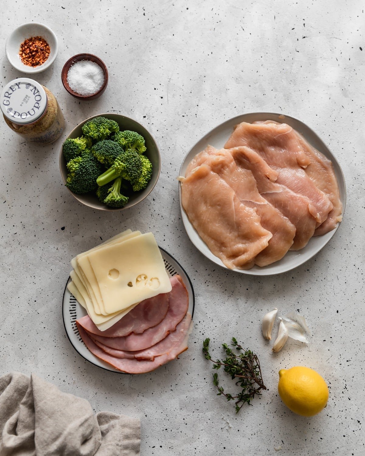 An overhead image of a white plate with raw chicken breasts on a grey table surrounded by a grey bowl of broccoli, white plate with ham and cheese, herbs, and spices.