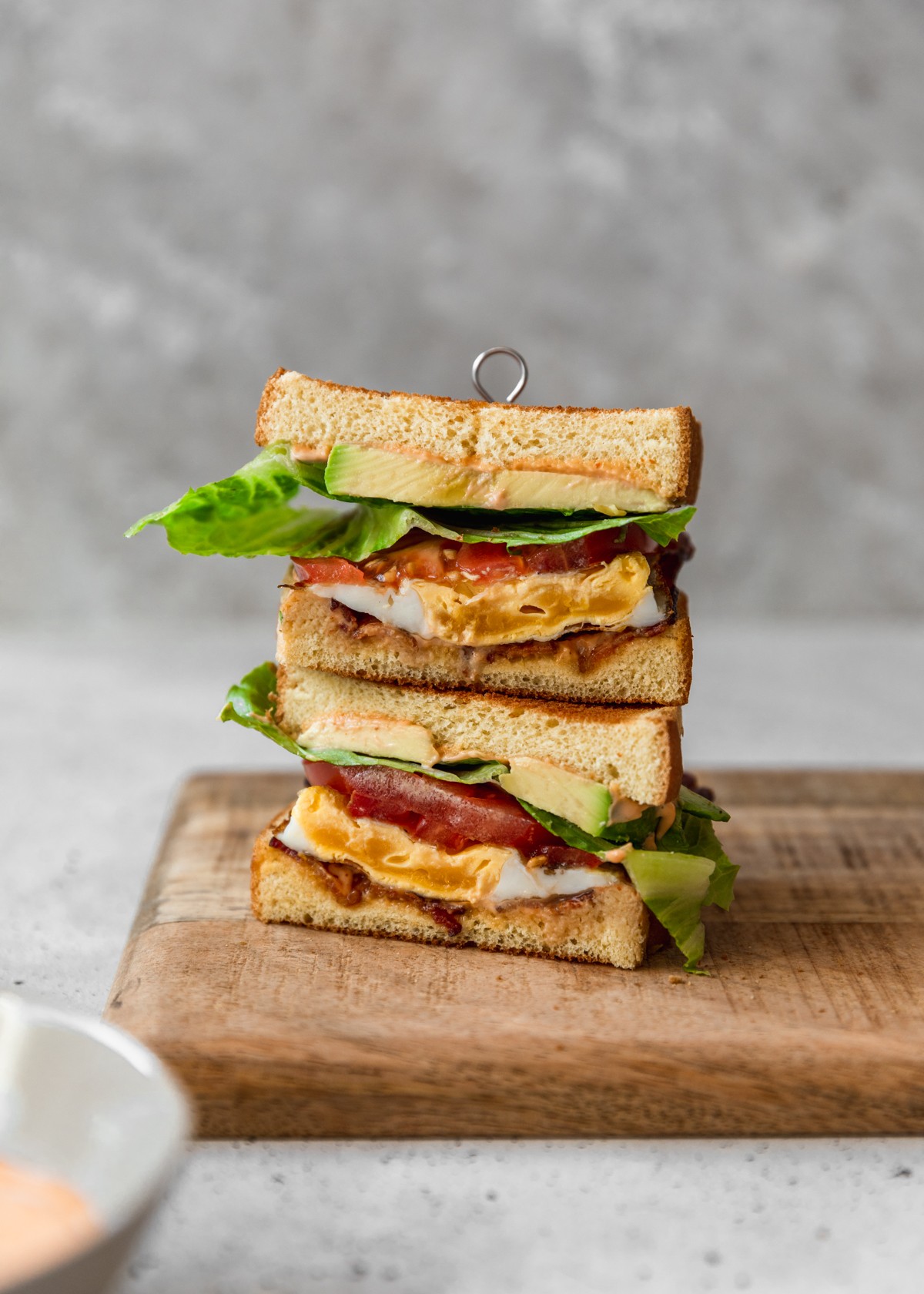 A side image of two breakfast BLT halves stacked on a wood board with a grey background.