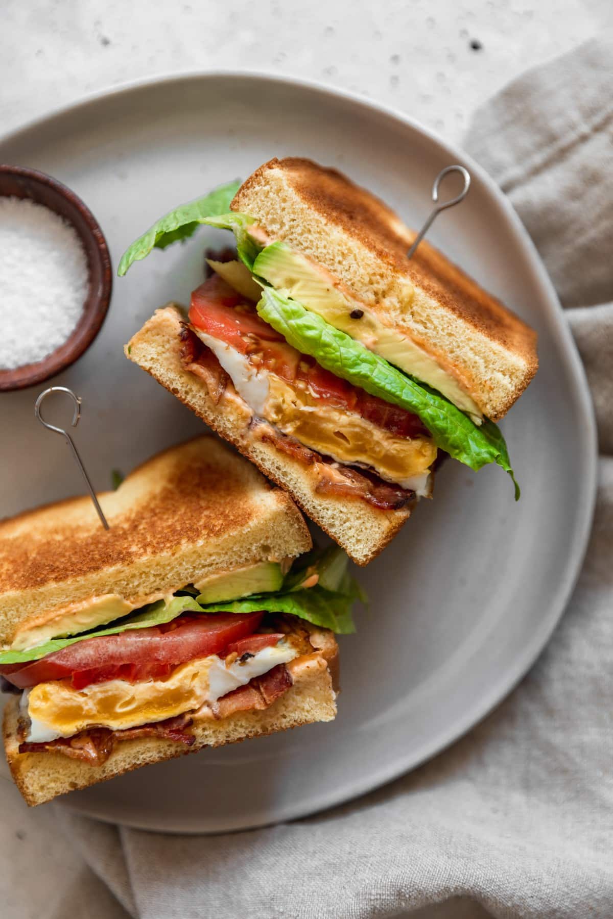 A closeup overhead image of a breakfast BLT sandwich with bacon, lettuce, tomatoes, avocado, and eggs, cut in half on a white plate.