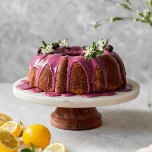 A side image of a pound cake with pink glaze on a marble cake stand with a grey background and lemons to the side.