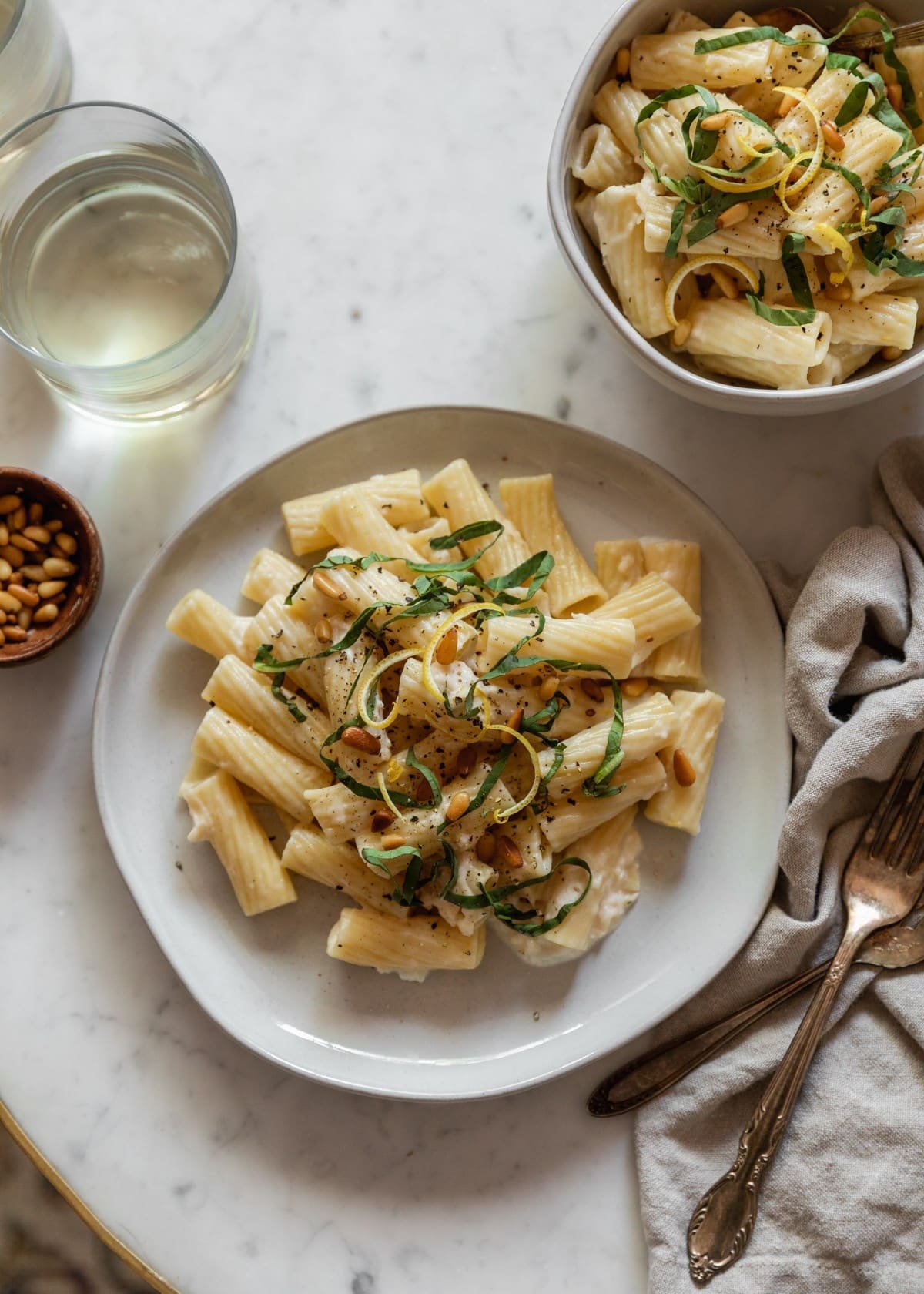 A closeup overhead image of a white plate topped with creamy lemon pasta on a marble counter next to a beige linen, vintage forks, a bowl of pasta, and a glass of white wine.