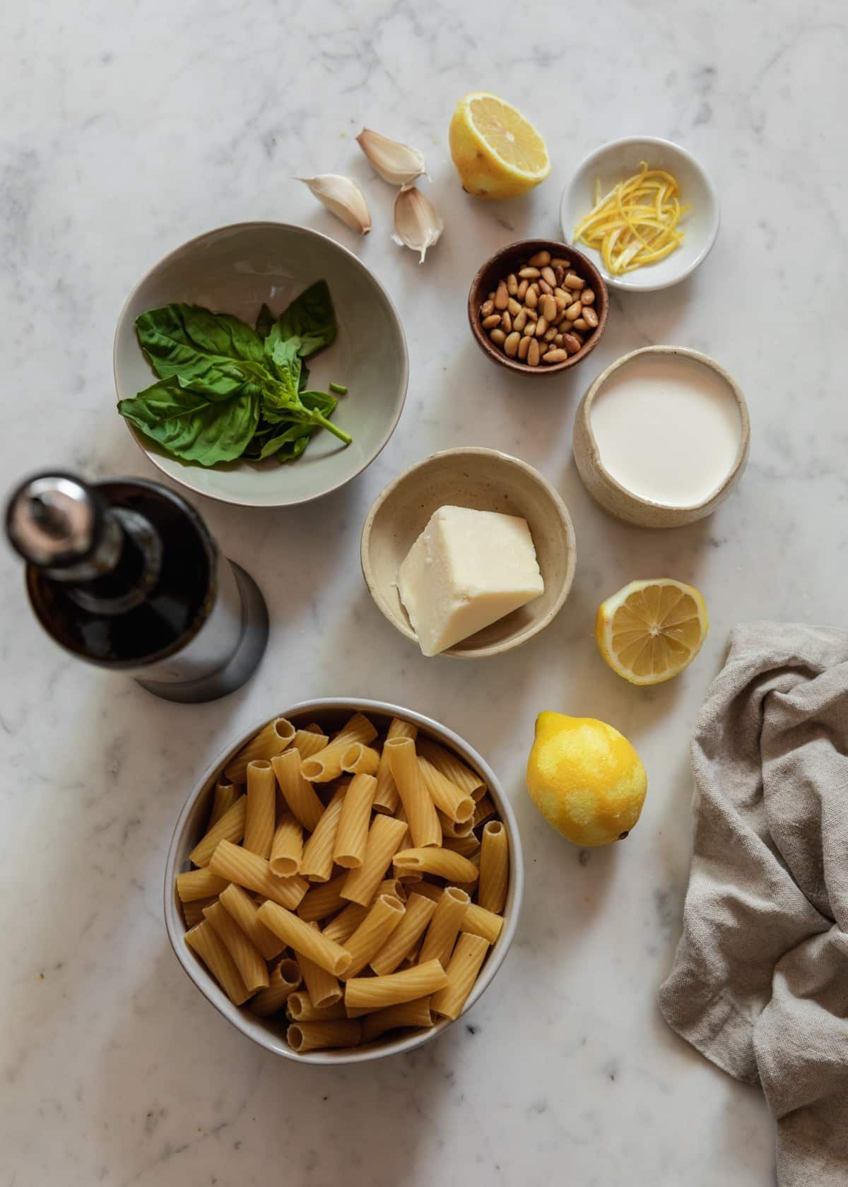 An overhead image of various white and grey bowls filled with rigatoni, Parmesan, cream, and basil on a marble counter next to lemons and a bottle of olive oil.