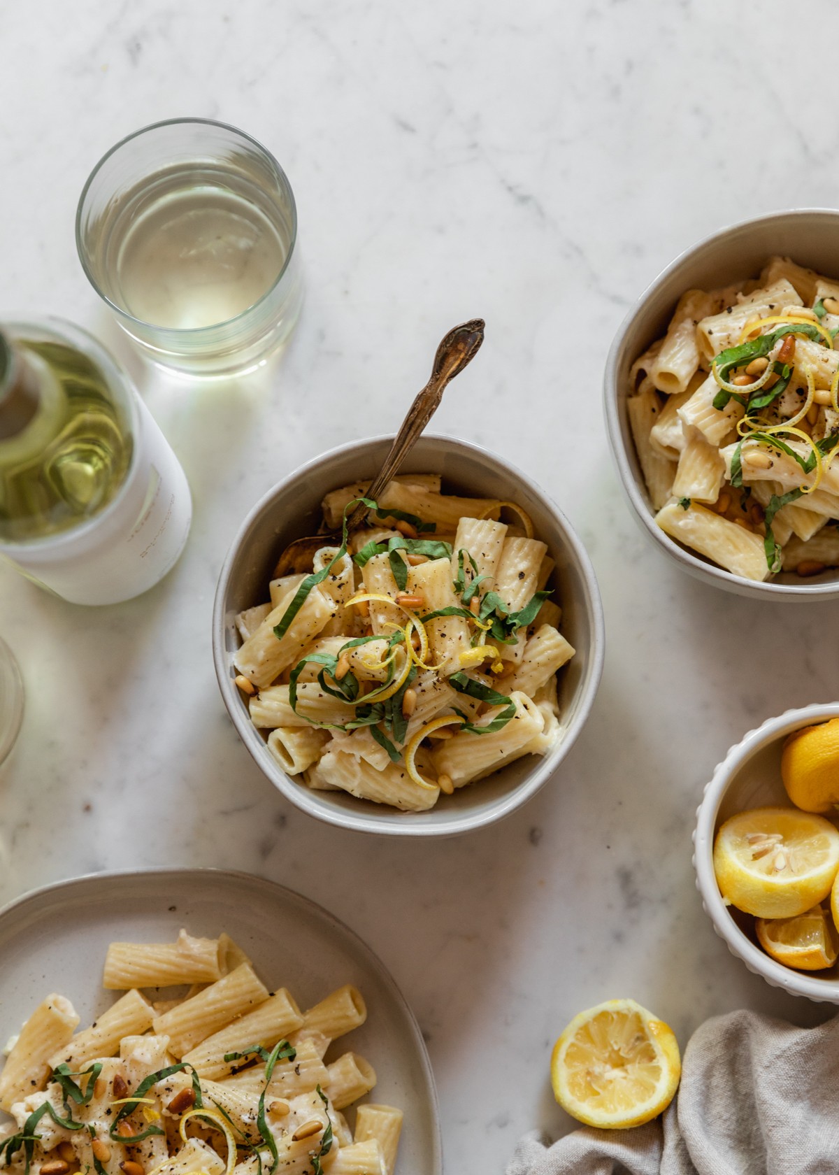 An overhead image of two bowls and a plate filled with creamy lemon pasta on a marble counter next to a bottle of white wine, a bowl of lemons, and a tan linen.