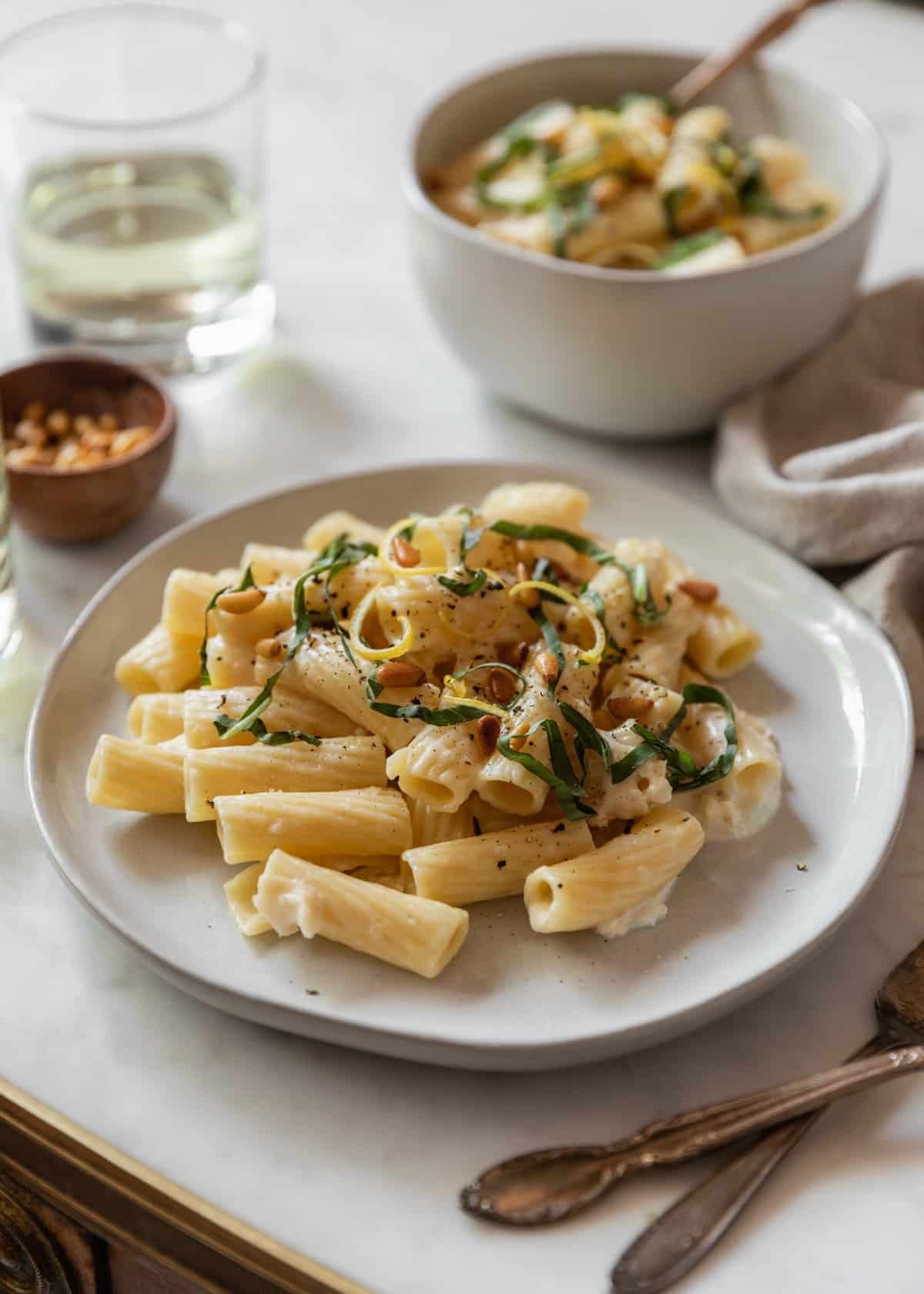 A closeup side image of a white plate of pasta with cream sauce, pine nuts, and basil on a marble counter next to a grey linen, wood bowl, and white bowl of pasta.