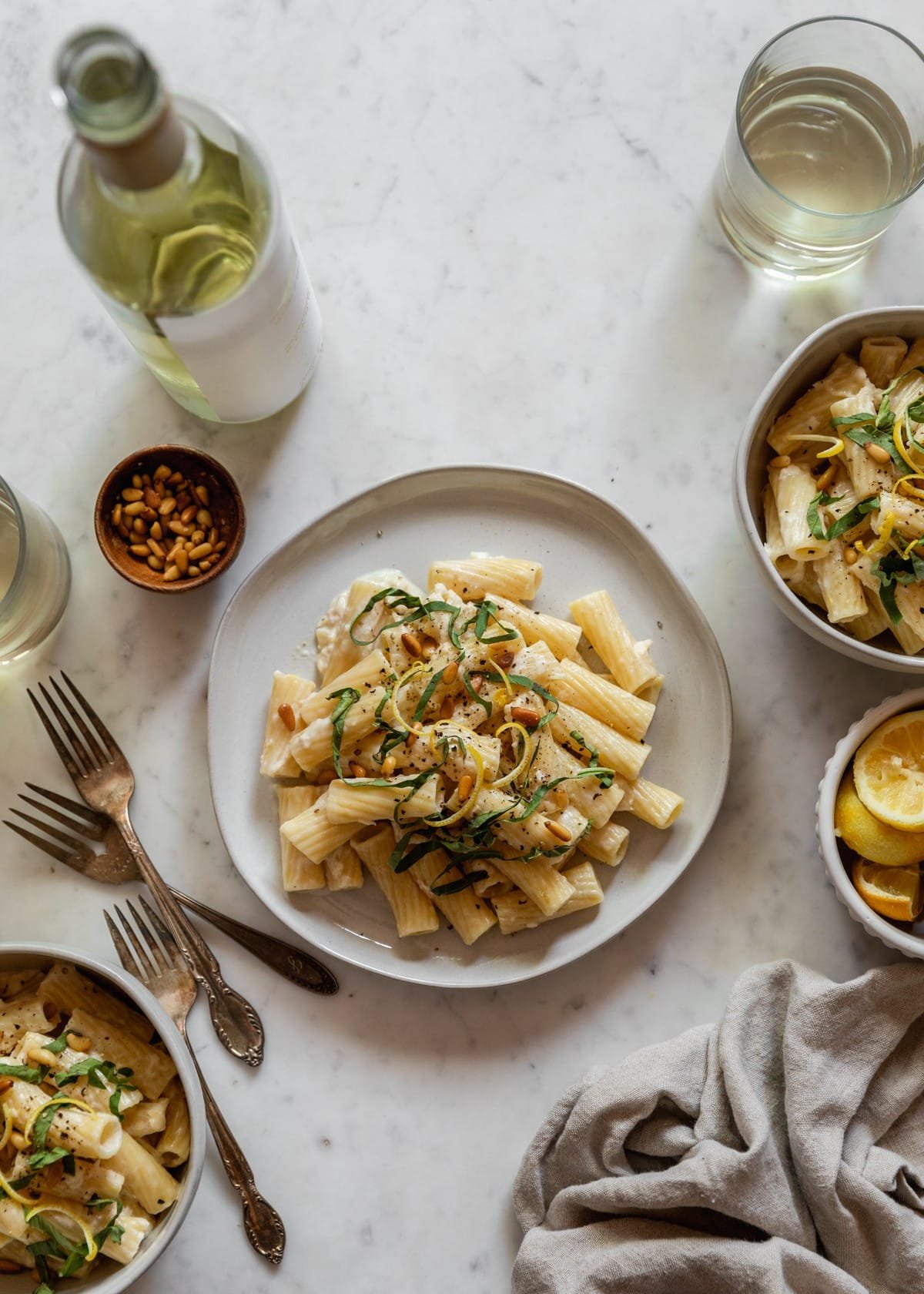 An overhead image of a plate and two bowls with creamy lemon pasta on a marble table next to a bowl of lemons, glasses of white wine, a wood bowl with pine nuts, forks, and a tan napkin.