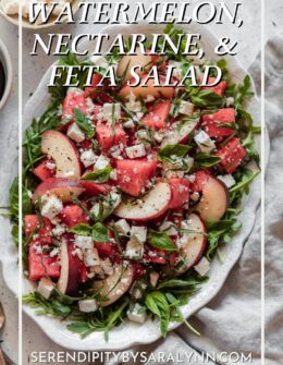 A closeup overhead image of a white oval dish with watermelon salad topped with feta, nectarines, arugula, and balsamic on a white linen with a white speckled background.