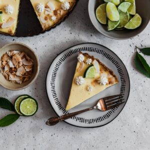 A closeup image of a slice of coconut key lime tart on a white plate placed on a grey table next to a bowl of limes and bowl of toasted coconut.