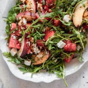 A closeup photo of a white plate of watermelon salad with arugula, feta, nectarines, and balsamic.