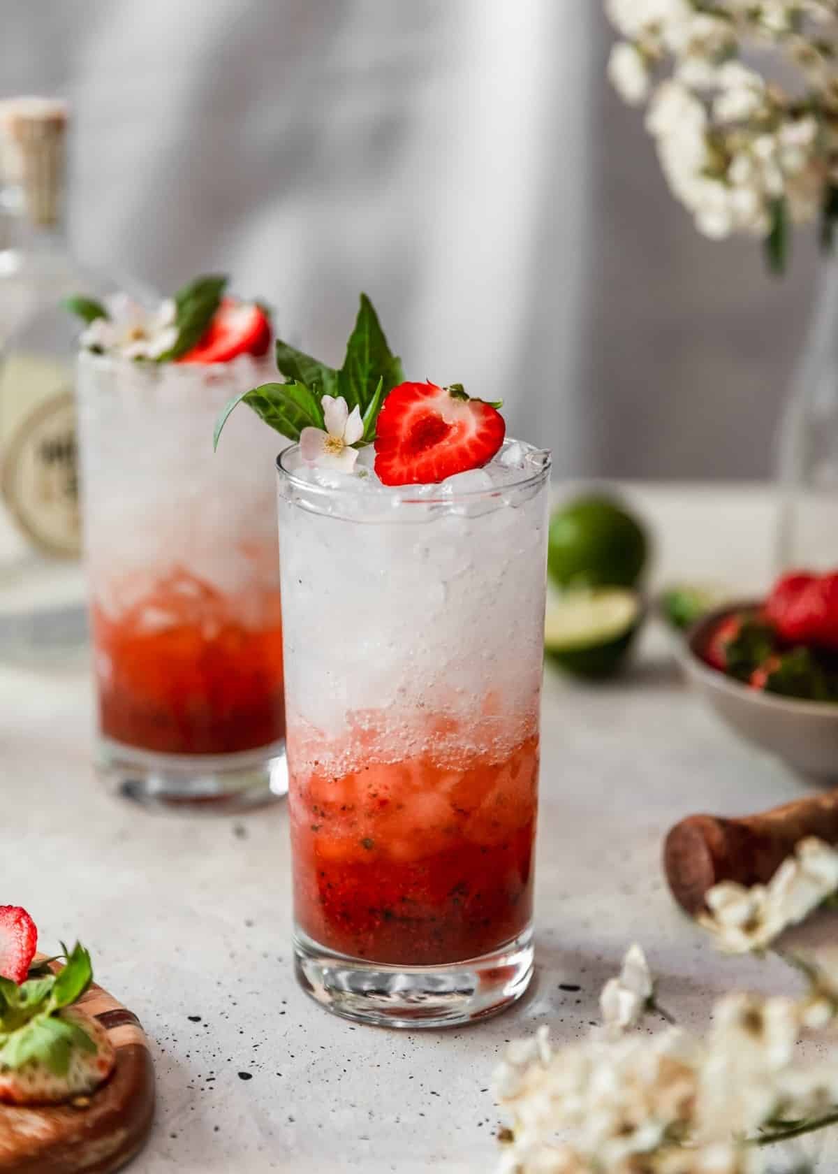 A tall clear glass filled  with a strawberry gin smash cocktail and garnished with a half strawberry, white flower and basil on a white table. In the background is another cocktail, a bottle of gin, a grey bowl of strawberries, a lime, and white blossoms.