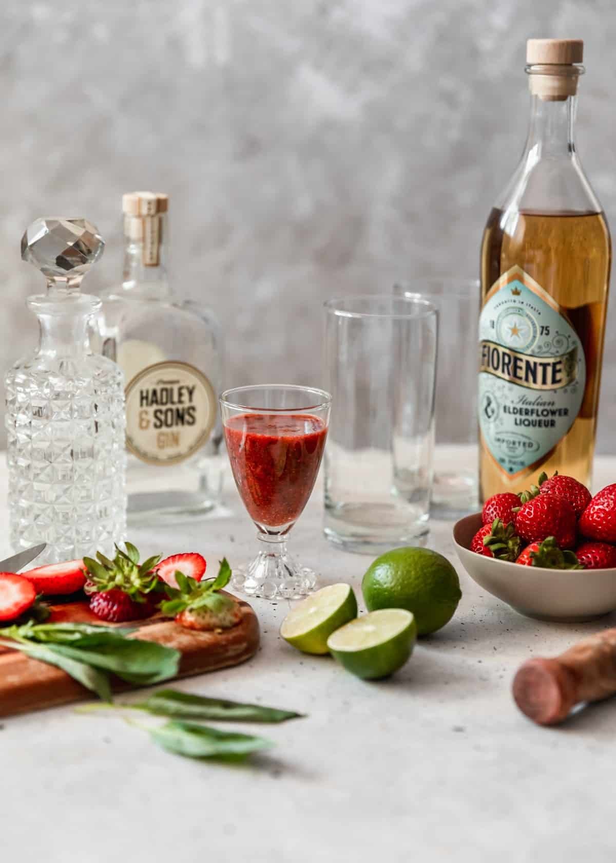 A glass of red juice, bowl of berries, alcohol bottles, a wood muddler, limes, and highball glasses on a white speckled counter.