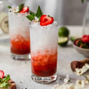 A side image of two strawberry cocktails on a white counter next to a bowl of berries, lime wedges, white blossoms, and a wooden muddler.