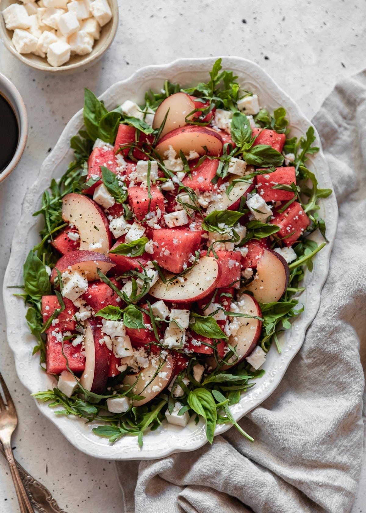 A closeup overhead image of a white dish of melon with arugula, feta, peaches, and basil on a white speckled counter next to a grey linen and vintage forks.