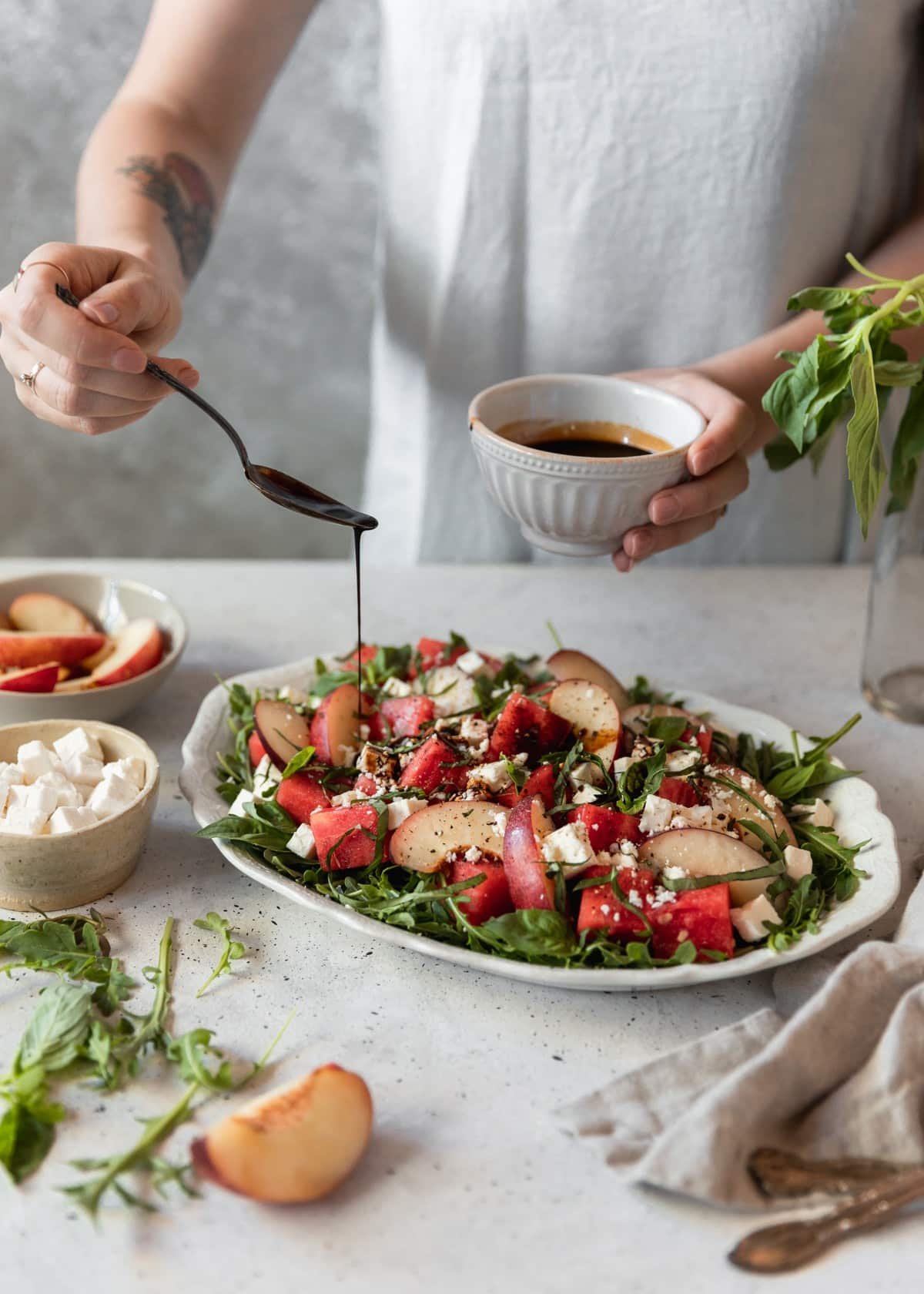 A woman wearing a white linen dress drizzling balsamic vinegar over watermelon salad with feta, nectarines, and arugula on a grey table next to a grey linen, cream bowl of feta cheese, and grey bowl of nectarines.