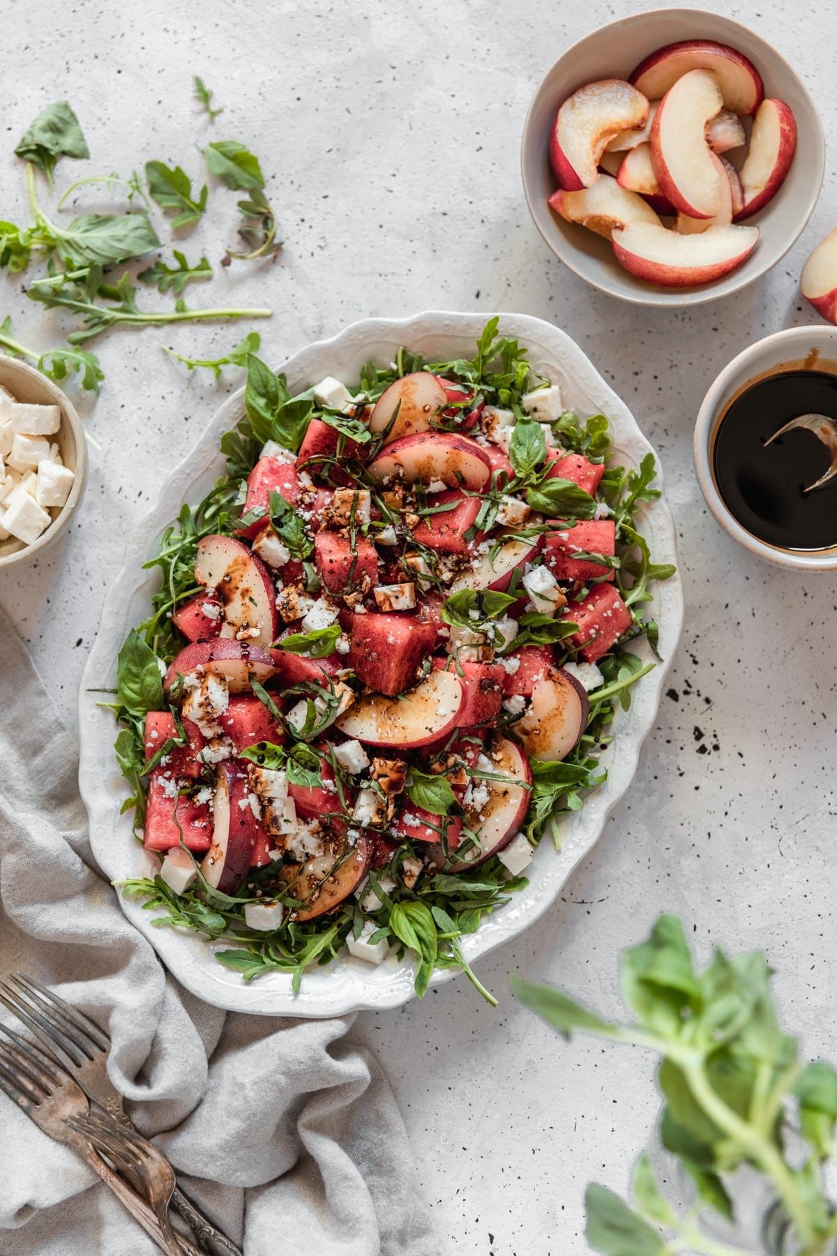 An overhead photo of an oval white tray of watermelon salad with feta cheese, greens, and peaches on a gray table next to a grey linen, white bowl of balsamic vinegar, a white bowl of feta, and a grey bowl of peaches.