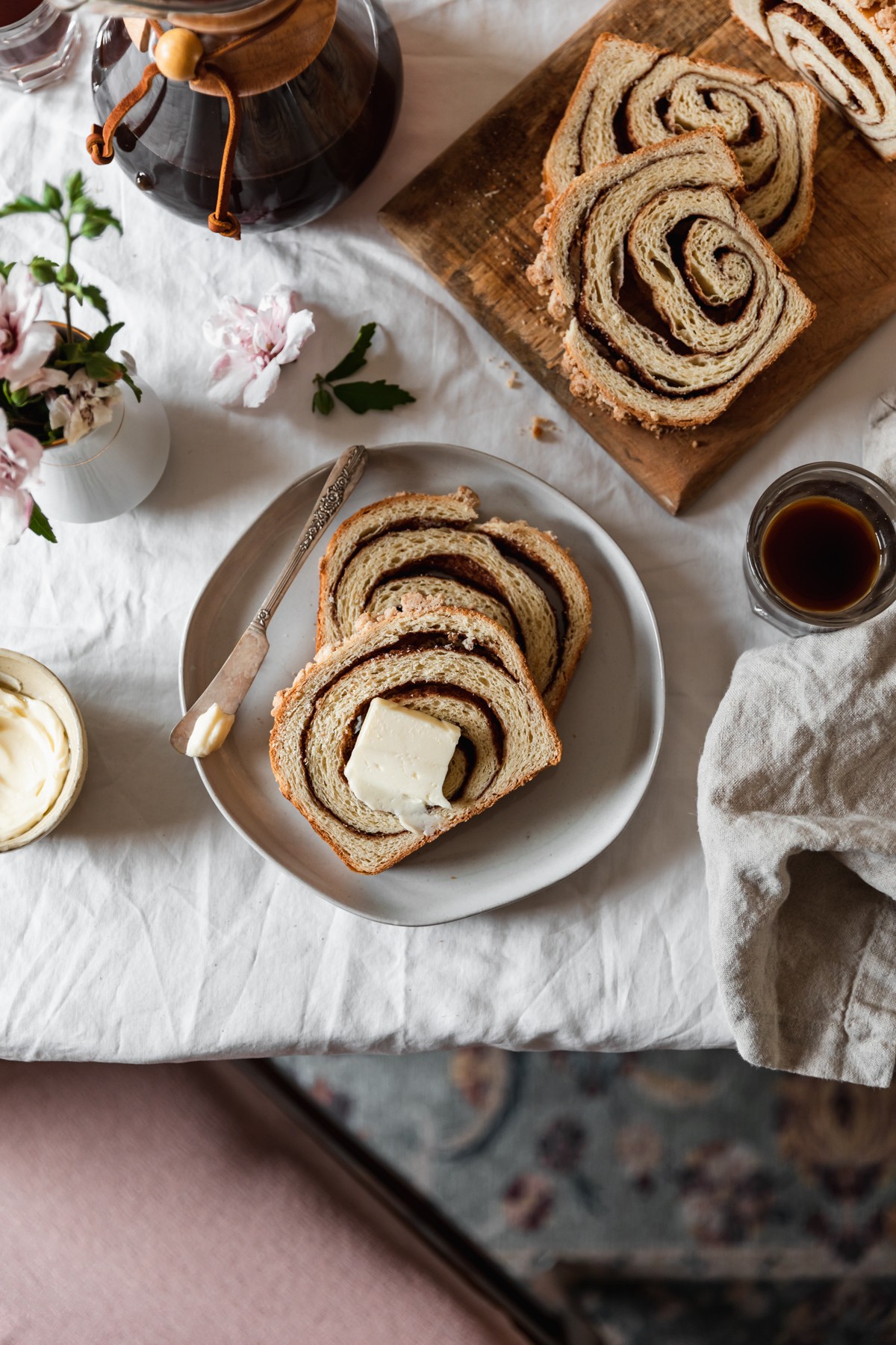 An overhead image of two slices of cinnamon swirl bread with streusel and a butter knife on a white plate. The plate is next to a wood board in the right corner topped with more bread, a beige linen, cups of coffee, a Chemex, and a bowl of butter on a white table cloth. In the bottom of the photo, you can see a green and pink vintage rug and a pink seat cushion.