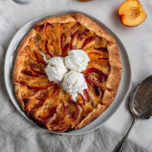 An overhead picture of a nectarine galette topped with three scoops of ice cream on a marble counter next to a metal pie server, beige linen, and half a sliced nectarine.