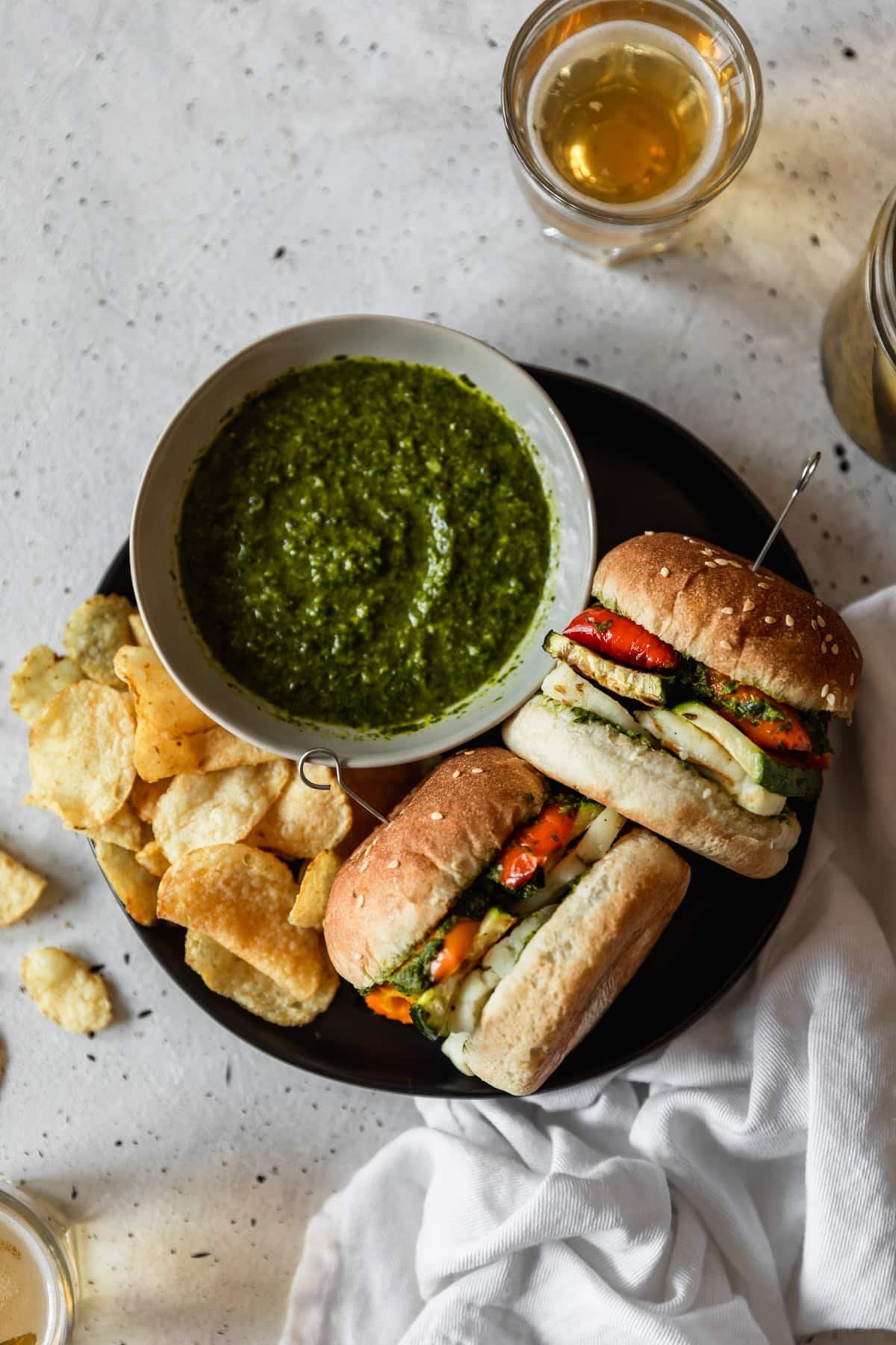 An overhead photo of a black plate with two halloumi and vegetable sandwiches, a grey bowl of herb sauce, and a handful of potato chips on a grey table next to a white linen, jar of pickles, and two cups of beer.