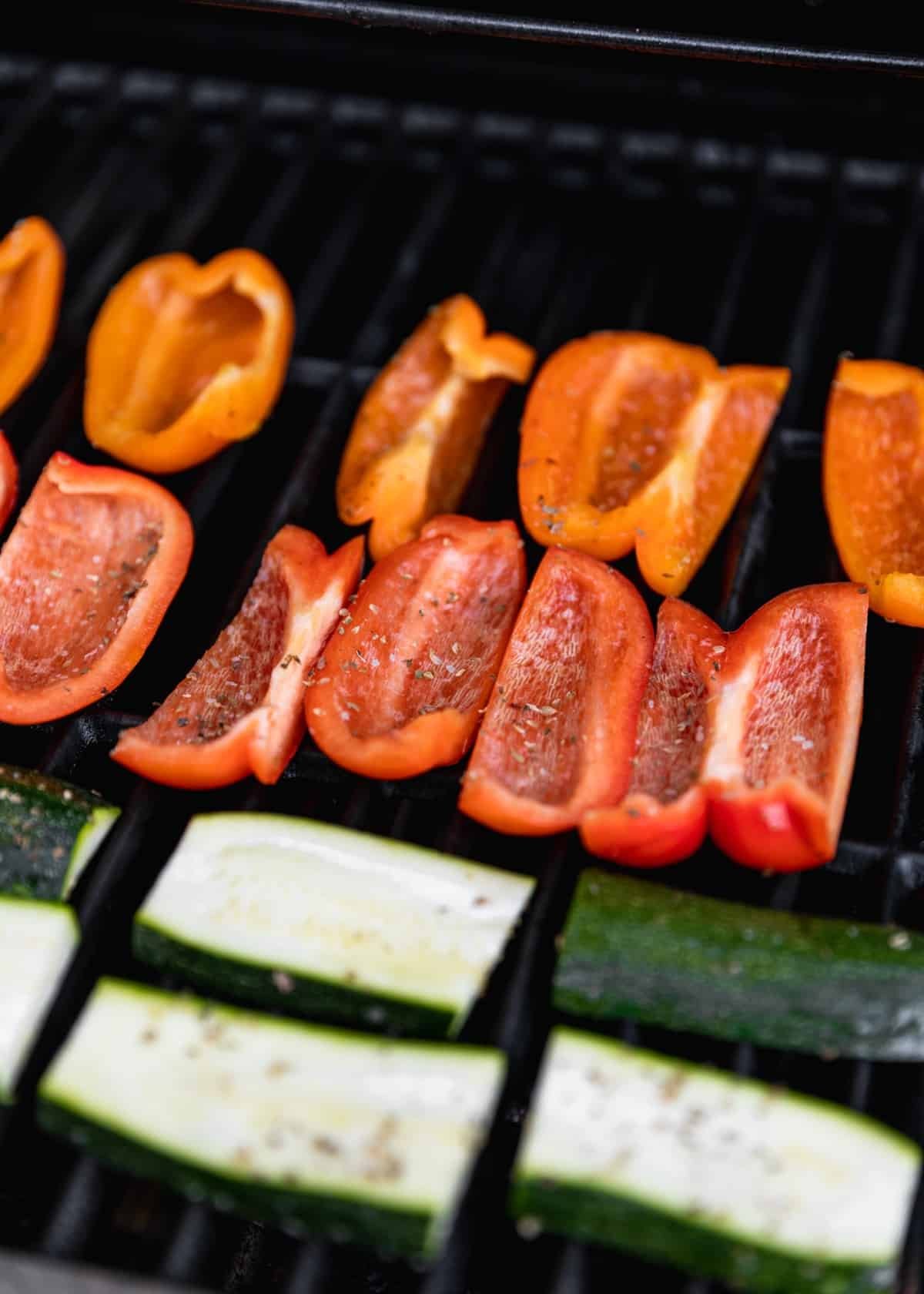 A side image of red and orage pepper slices and zucchini slices cooking on a grill.