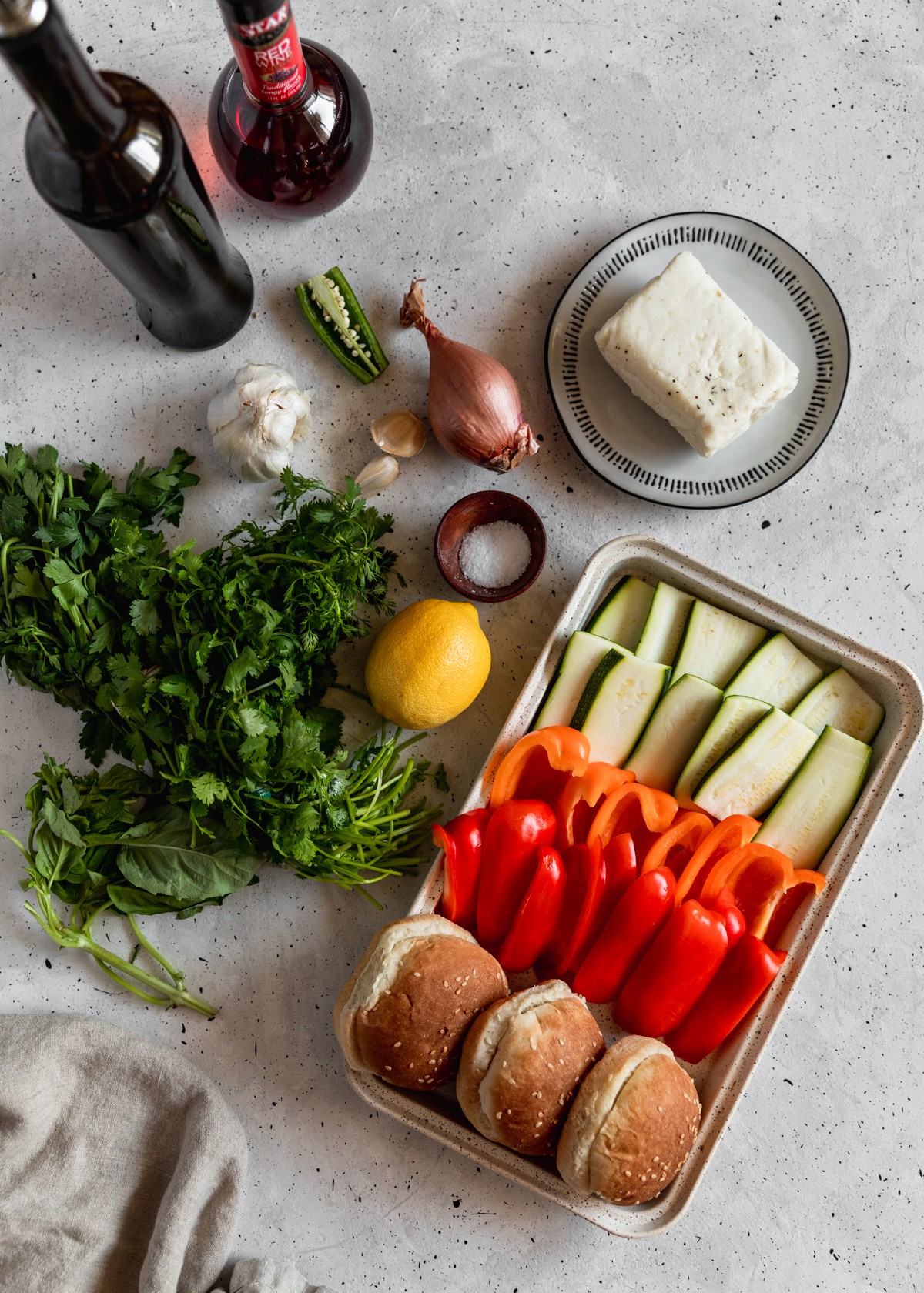 A white tray with peppers, zucchini slices, and buns next to a beige linen, bunches of herbs, a lemon, a shallot, garlic, a white plate of halloumi, a bottle of olive oil, and a bottle of red wine vinegar on a grey table.