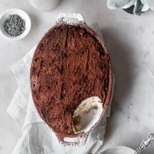 A closeup image of a white oval tray of tiramisu with a scoop taken out of it on a white marble counter next to greenery and a white dish of dried lavender.