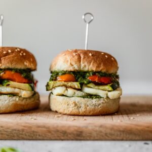 A closeup side image of a halloumi sandwich with veggies and green sauce on a wood board with a light grey background.