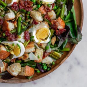 A closeup overhead image of a panzanella salad with greens, spring veggies, and eggs on a marble counter.