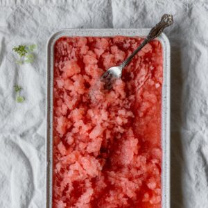 A closeup overhead image of half a white tray of pink granita with a fork on top placed on a white linen.