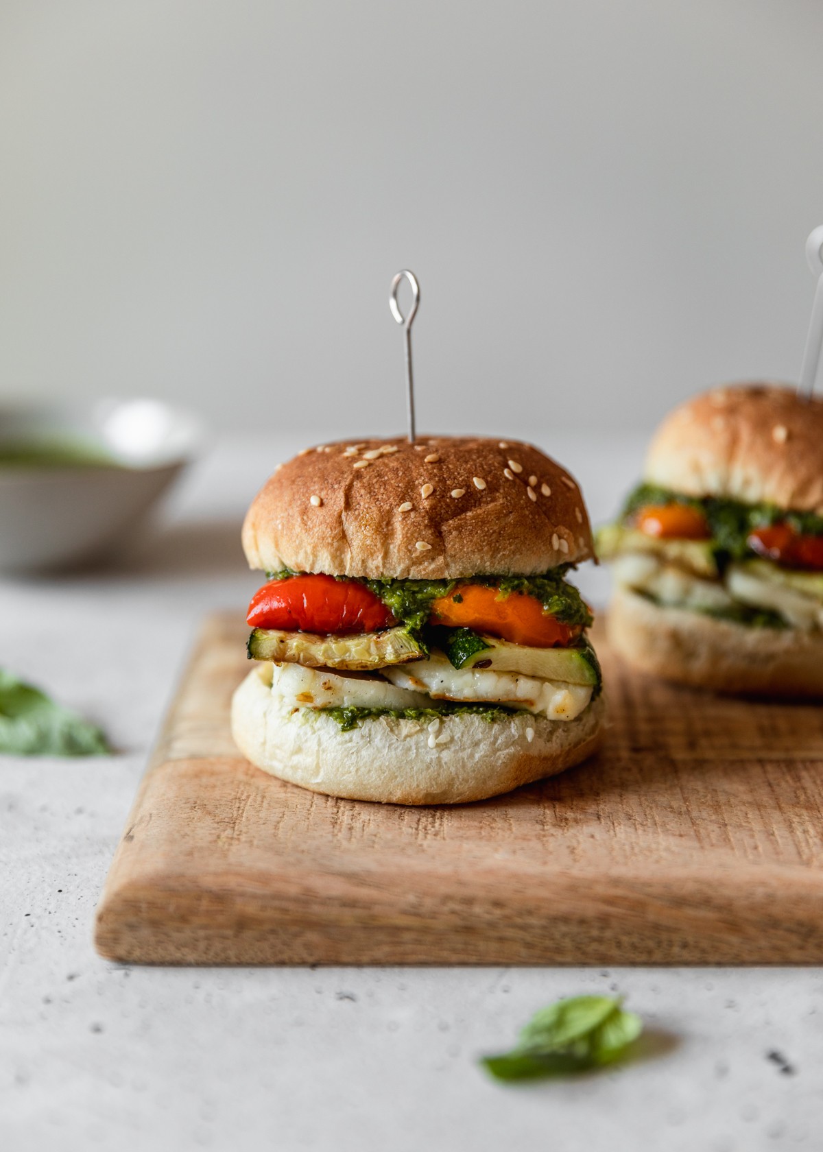 A side image of two vegetarian burgers on a wood board next to basil leaves and a grey bowl of green sauce on a white speckled table.
