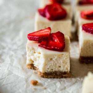 A closeup image of a strawberry cheesecake bar with a bite taken out of it on a parchment paper with more bars in the background.