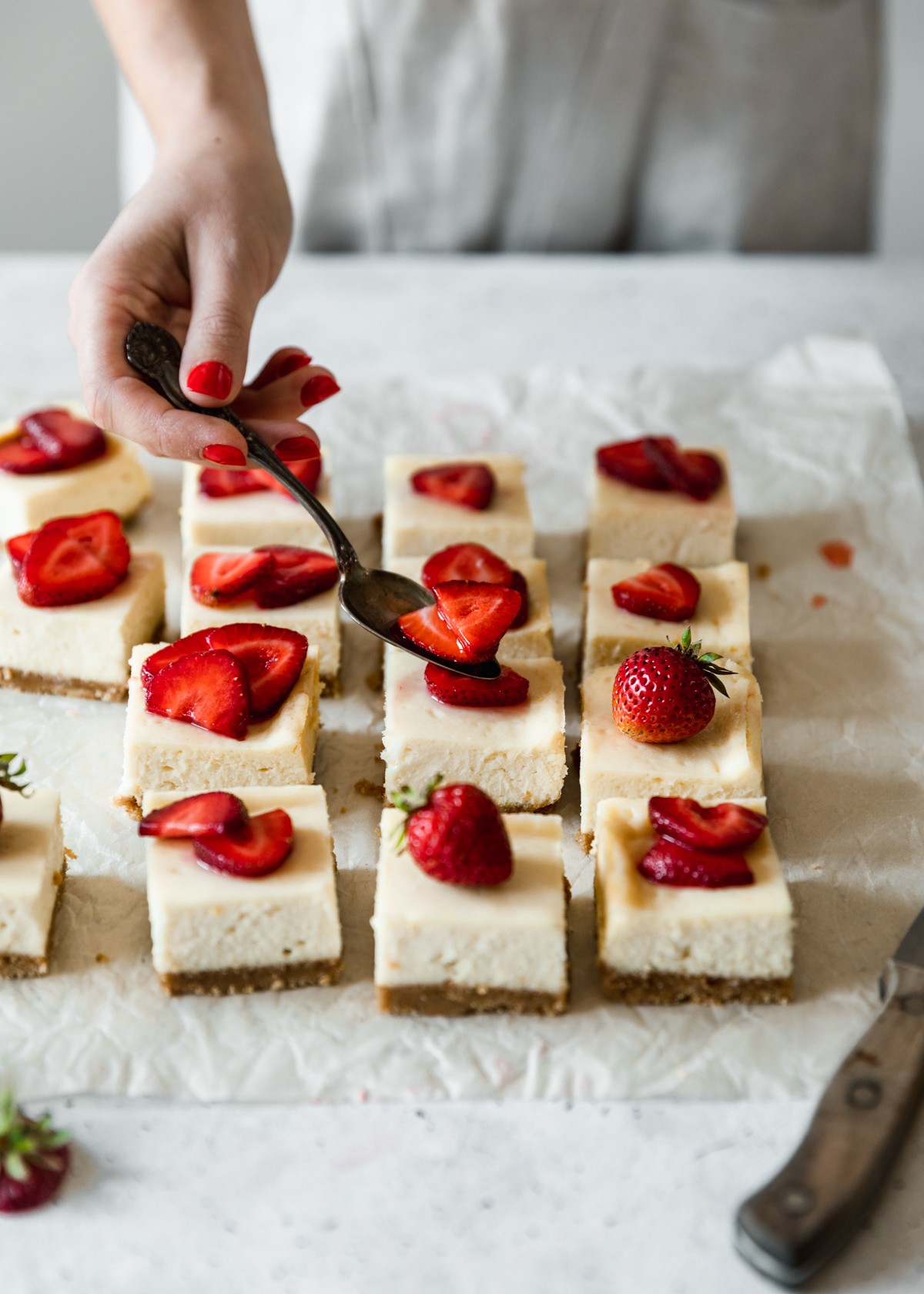 Rows of strawberry cheesecake bars on white parchment paper placed on a white counter. In the background, a woman with red finger nails wearing a beige apron is spooning strawberries over one of the middle bars.