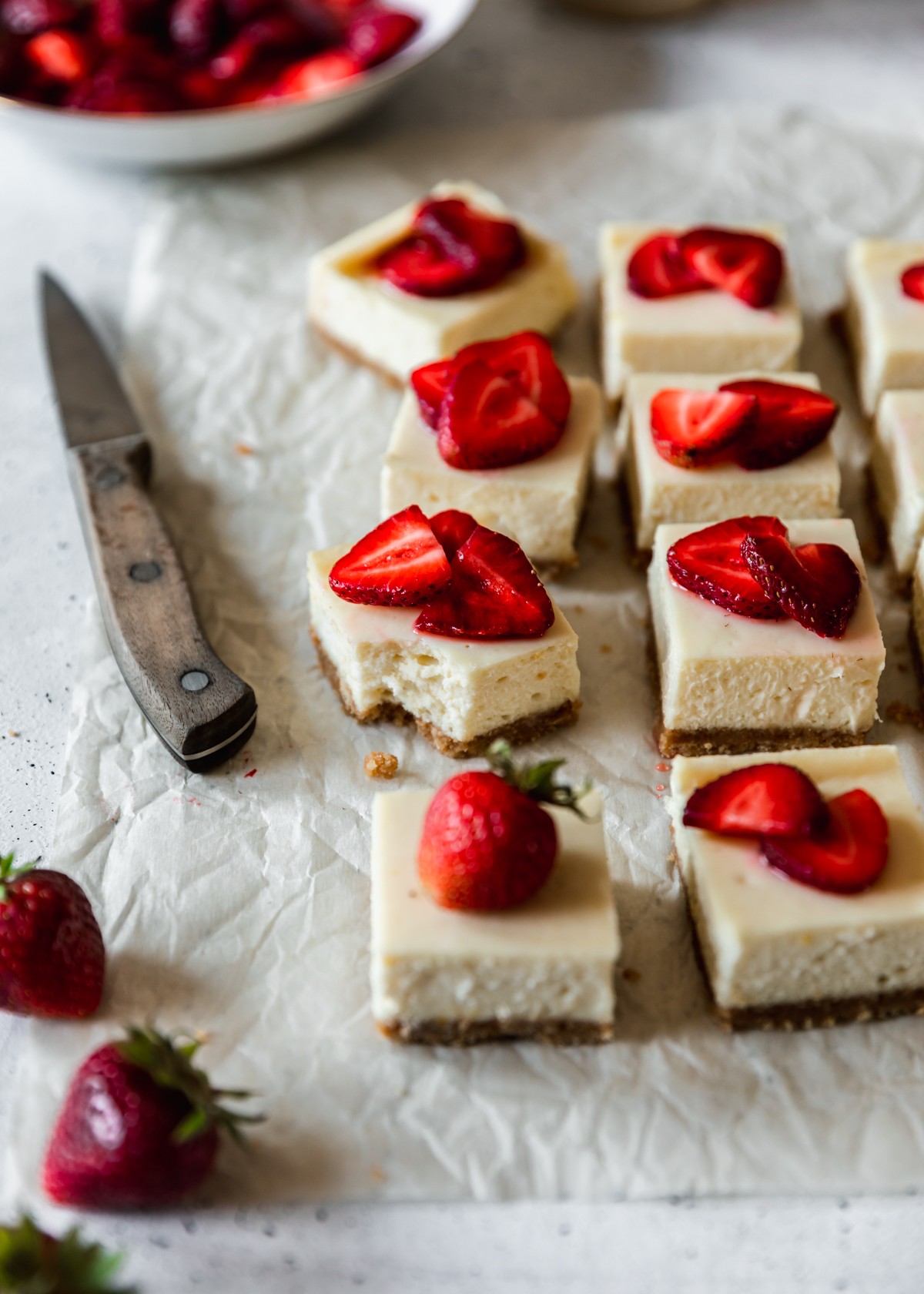Two rows of strawberry cheesecake bars on white parchment paper next to a wooden knife and white bowl of strawberries on a white speckled table. The second bar on the left has a bite taken out of it.