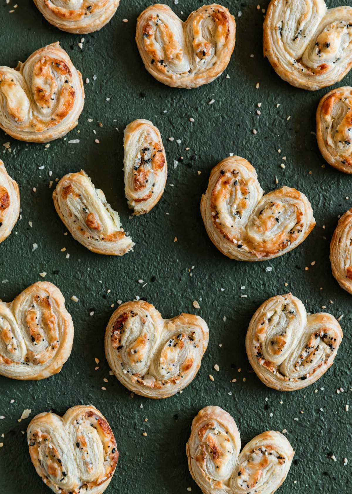 A close up overhead image of savory palmiers with cream cheese, Parmesan, and everything bagel spice on a dark green table. In the left upper corner, one of the palmiers is broken.