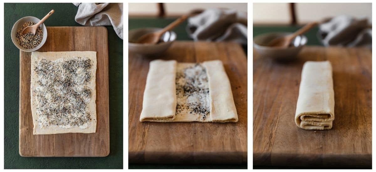 Three images; on the left, an overhead of a wood board with puff pastry coated in cream cheese and seeds on a green table next to a beige linen. In the middle, a closeup of partly folded puff pastry on a wood board with a grey bowl and beige linen in the background. On the right, the puff pastry is folded on top of each other.