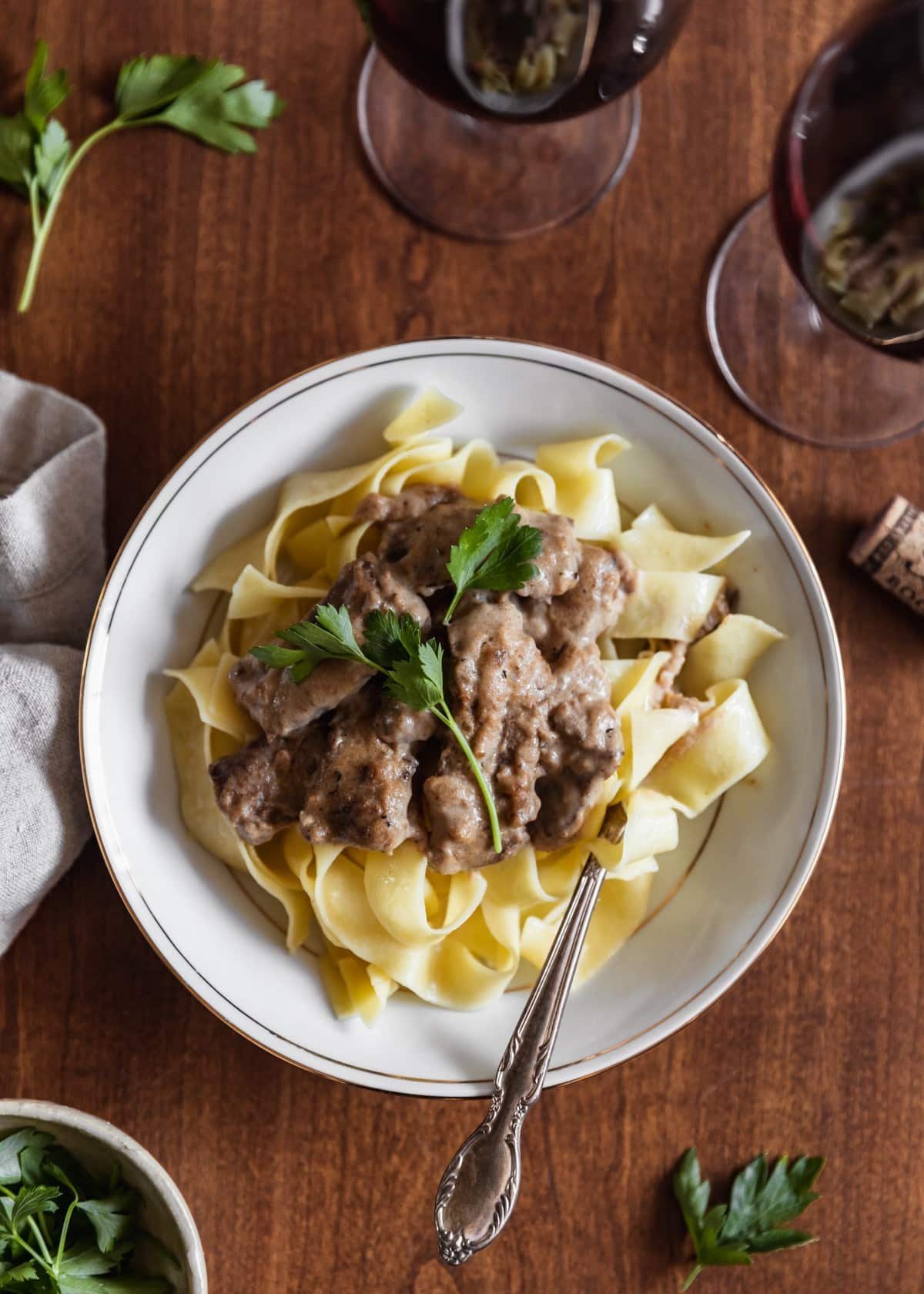 A close up overhead image of a white bowl of beef stroganoff and pasta with a vintage fork on a wood table. Next to the bowl is a beige linen, glass of wine, wine cork, and white bowl of parsley.