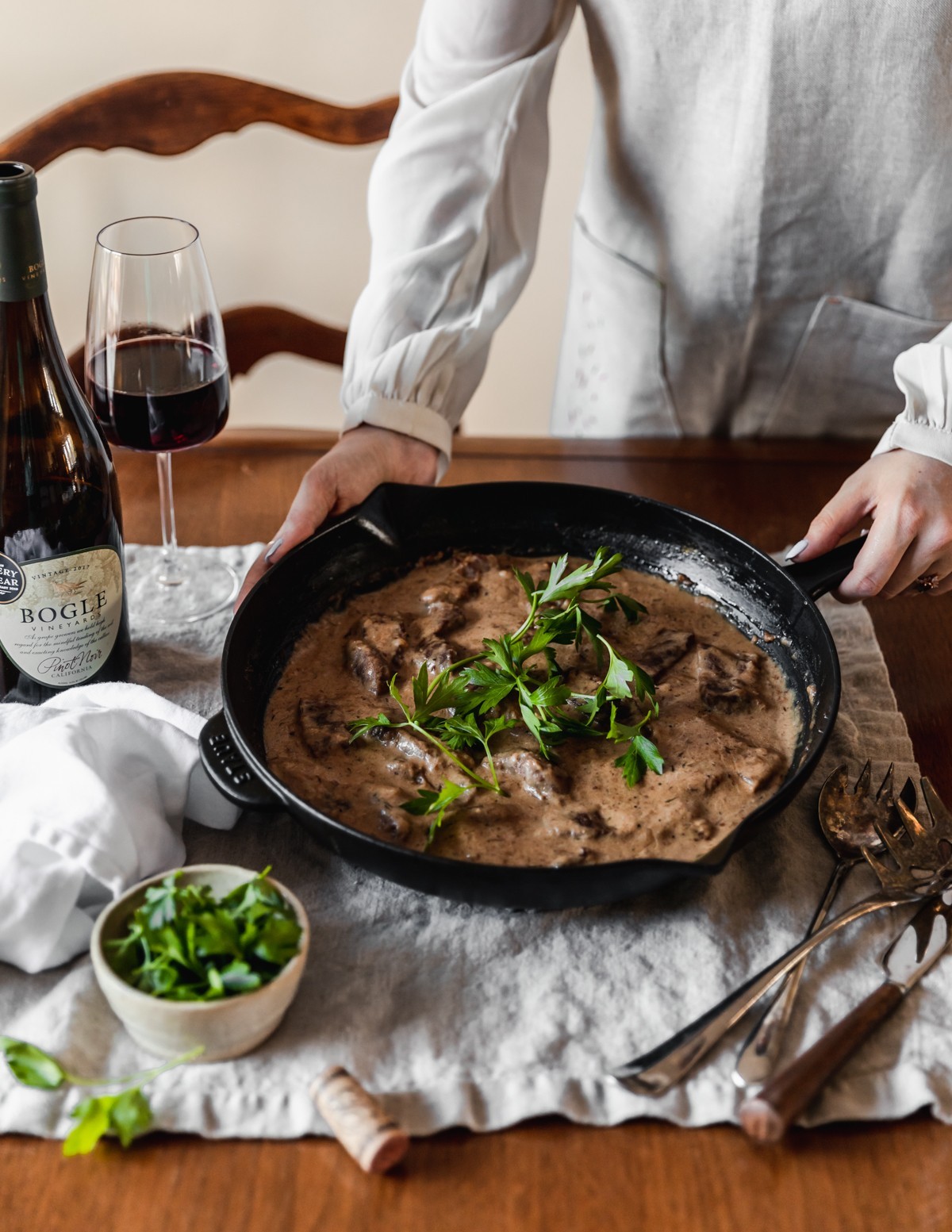 A side image of a woman wearing a white flowly long sleeve shirt and beige linen apron holding a black cast iron pan of beef stroganoff with shallots and brandy garnished with parsley. The pan is on a beige linen on a wood table next to vintage utensils, a white bowl of parsley, a white linen, a bottle of wine, and a glass of red wine.