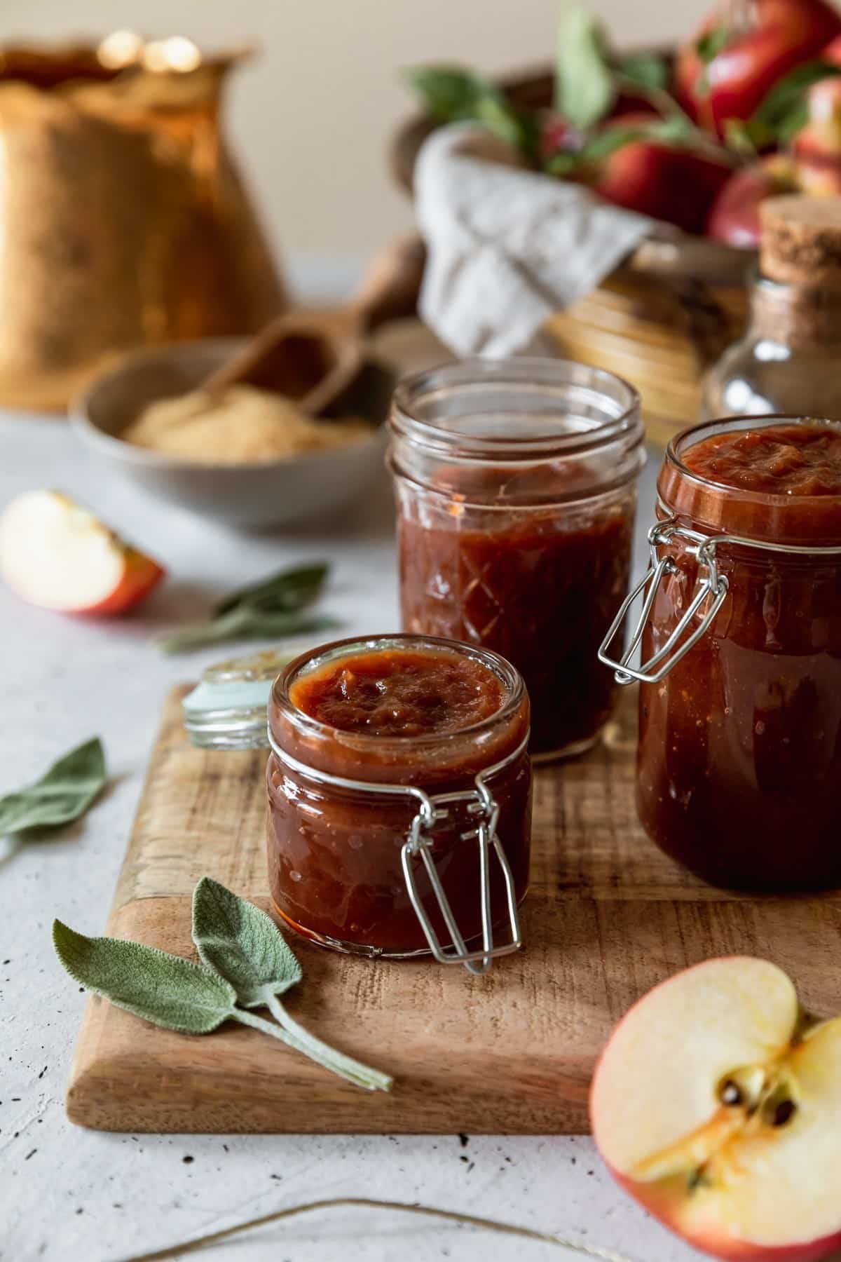 A closeup side image of a small jar of apple butter with two larger jars on the background on a wood board with sage leaves. The board is on a white speckled table with a gold teapot, basket of apples, and grey bowl of sugar in the background.