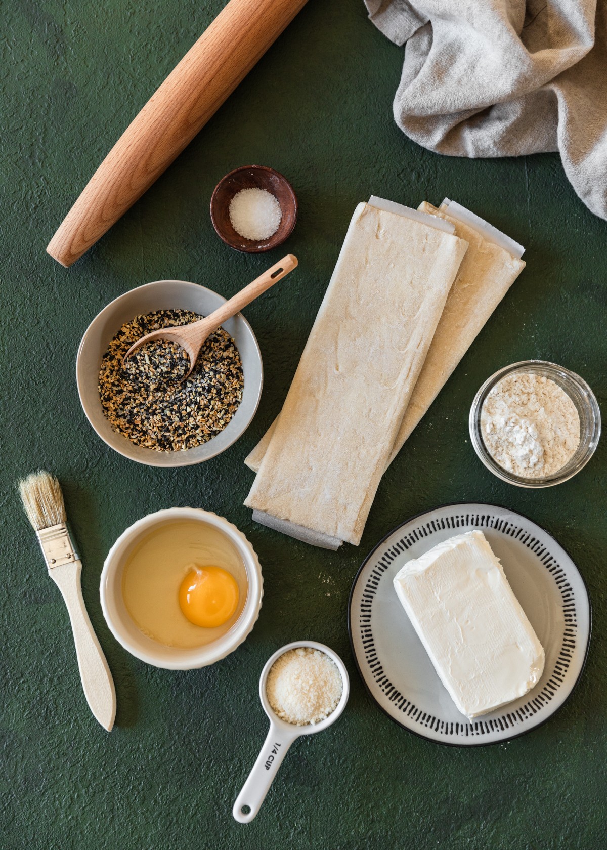 An overhead image of puff pastry, a jar of flour, a grey bowl of everything spice, a white plate with cream cheese, a white bowl with an egg, a rolling pin, and a pastry brush on an emerald green table.