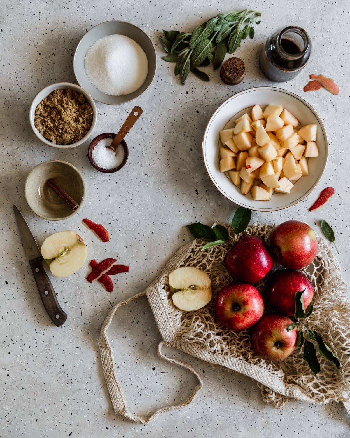 A mesh bag filled with apples on a grey counter next to a white bowl of diced apples, white and grey bowls with sugar, brown sugar, and cinnamon, a bunch of sage, and a bottle of vanilla extract.